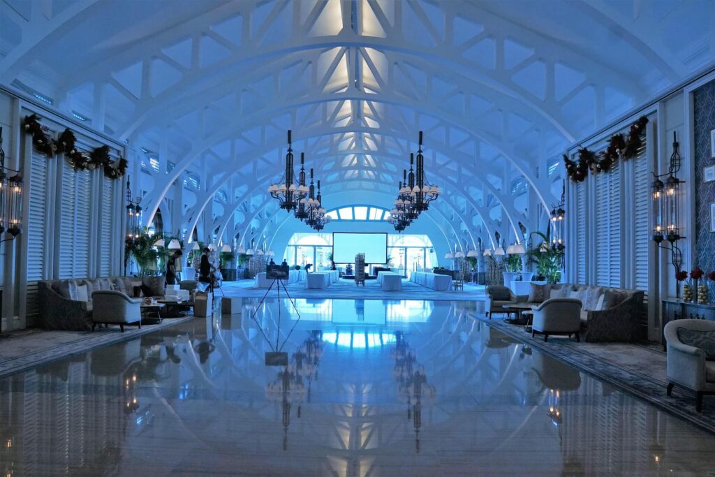 Spacious hotel lobby with high arched ceilings, chandeliers, and lounge seating reflected on a polished marble floor.